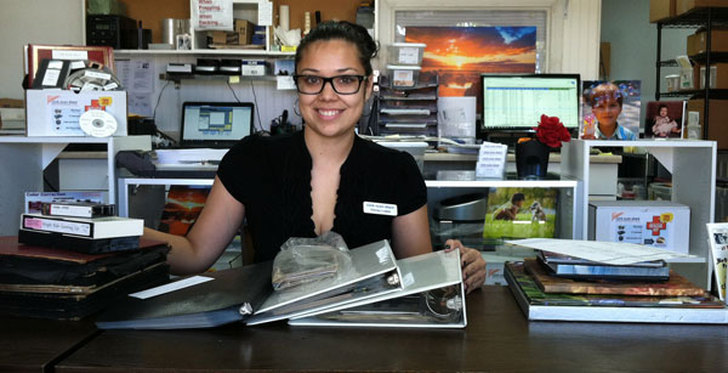 Behind a stack of binders and albums full of old photos, a helpful employee at Click Scan Share in San Diego assists new customer in starting a photo book project.